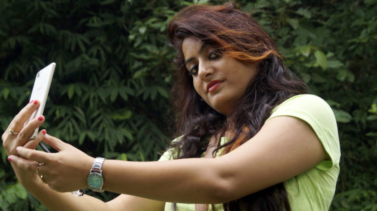 Vibrant woman taking a selfie outdoors with lush green foliage, showcasing lifestyle and nature photography.