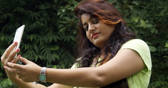 Vibrant woman taking a selfie outdoors with lush green foliage, showcasing lifestyle and nature photography.